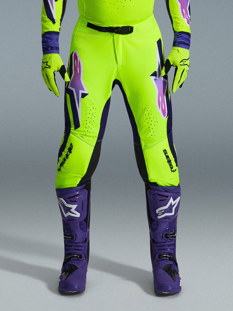 Alpinestars 2026 Supertech Vista broek, motorcrossbroek, fluorgeel, paars en zwart, gedragen met bijpassende handschoenen en paarse laarzen, voorzien van grote gradiëntlogo's op de dijen, geperforeerde zones voor ventilatie en een professioneel atletisch ontwerp voor off-road racen