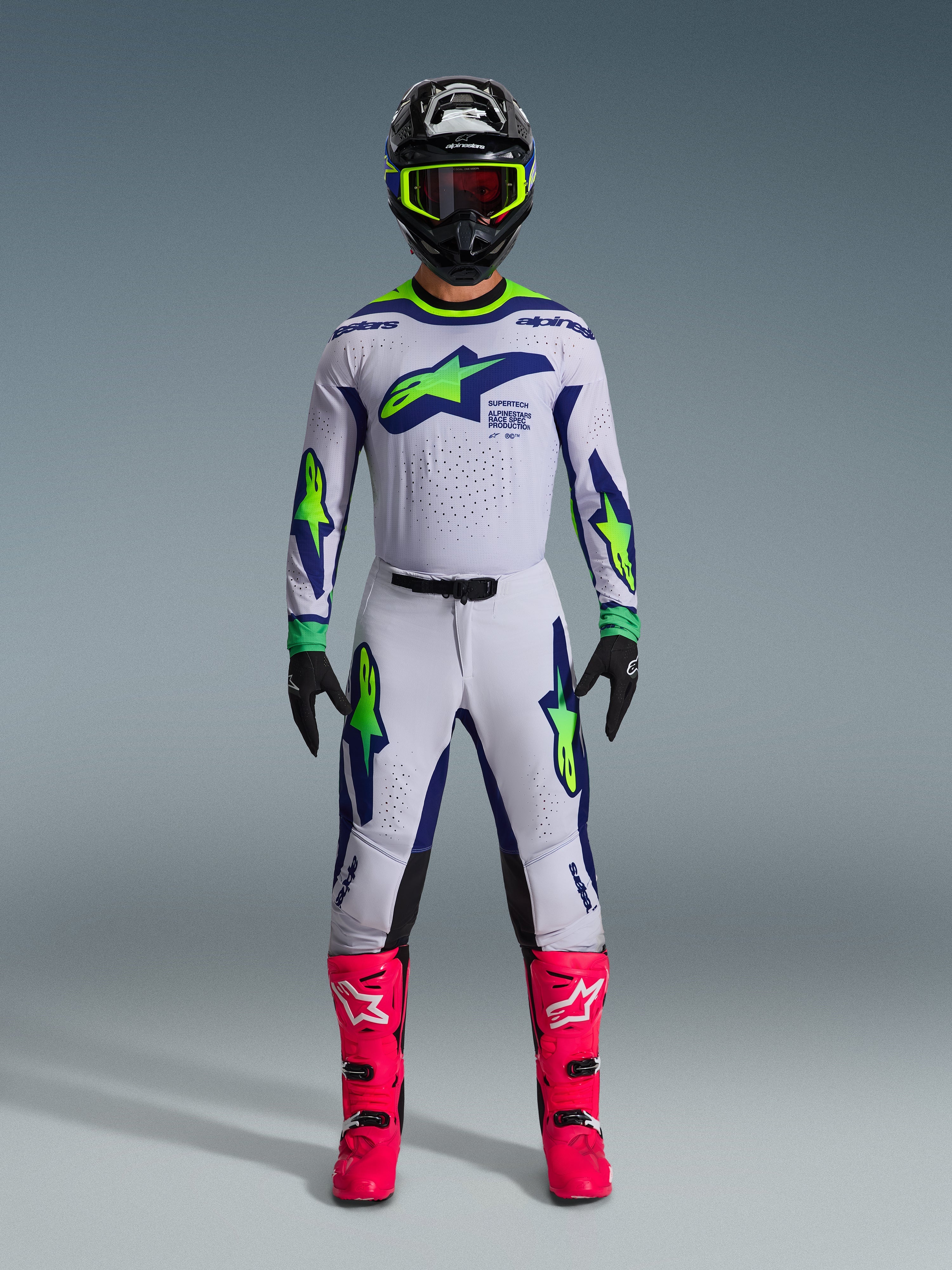 Alpinestars Supertech Vista Gear Set 2026, pilote de motocross portant une tenue complète, maillot et pantalon blancs avec accents bleus et vert fluo, casque noir avec finitions jaune fluo, bottes rose vif, équipement de course tout-terrain professionnel