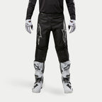 Alpinestars Pantalon Fluid Graphite 2024, pantalon de motocross, noir et argent, avec logos Alpinestars argentés sur les cuisses, porté avec des bottes et des gants de motocross blancs et noirs, design sportif pour la course tout-terrain