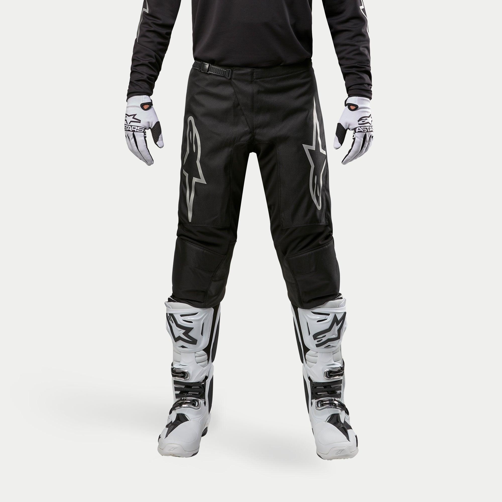 Alpinestars Pantalon Fluid Graphite 2024, pantalon de motocross, noir et argent, avec logos Alpinestars argentés sur les cuisses, porté avec des bottes et des gants de motocross blancs et noirs, design sportif pour la course tout-terrain