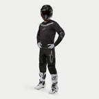Pantalon Alpinestars Fluid Graphite 2024, pantalon de motocross, noir et argent, porté avec un maillot noir assorti, un casque noir, des gants blancs et des bottes blanches, logos Alpinestars argentés sur les cuisses, coupe athlétique ergonomique pour la course tout-terrain.