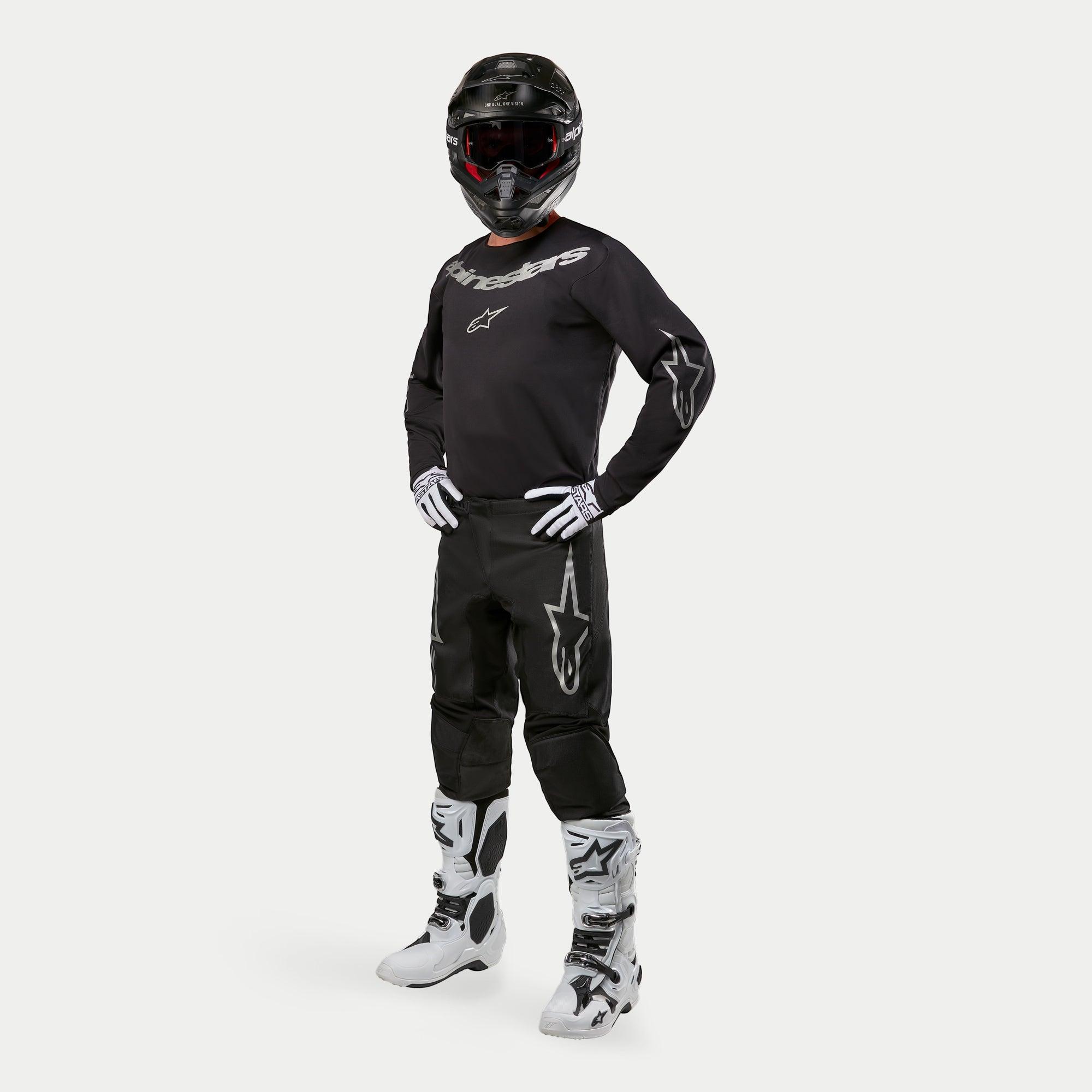 Pantalon Alpinestars Fluid Graphite 2024, pantalon de motocross, noir et argent, porté avec un maillot noir assorti, un casque noir, des gants blancs et des bottes blanches, logos Alpinestars argentés sur les cuisses, coupe athlétique ergonomique pour la course tout-terrain.