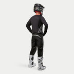 Pantalon Alpinestars Fluid Graphite 2024, pantalon de motocross, noir et argent, vue arrière d'un pilote portant un maillot noir, un casque noir et des bottes blanches et noires, avec logo argenté au bas du dos et une construction durable en poly-tissu pour la course tout-terrain