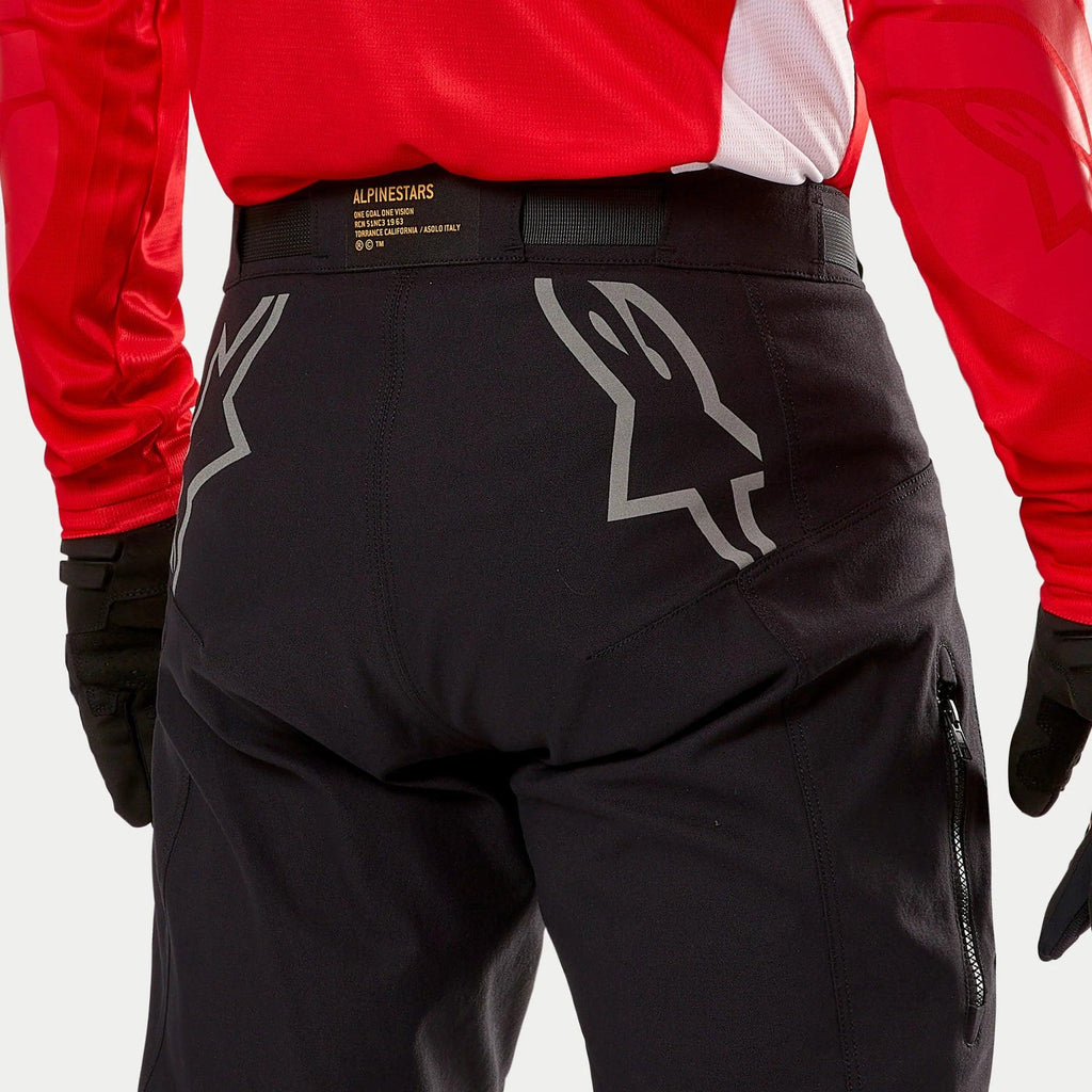 Alpinestars Techdura Pants, Black Enduro Pants, Rear view featuring large grey Astars logos on the seat, worn with a red jersey and black gloves, durable material with side zipper and branded waistband