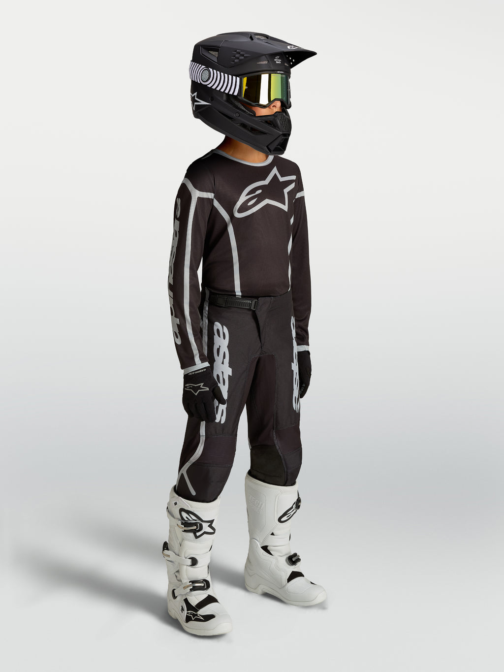 Alpinestars Jeugd Fluid Apex V2 broek, zwart en zilver motocrossuitrusting op model met jersey, helm en witte motorlaarzen.