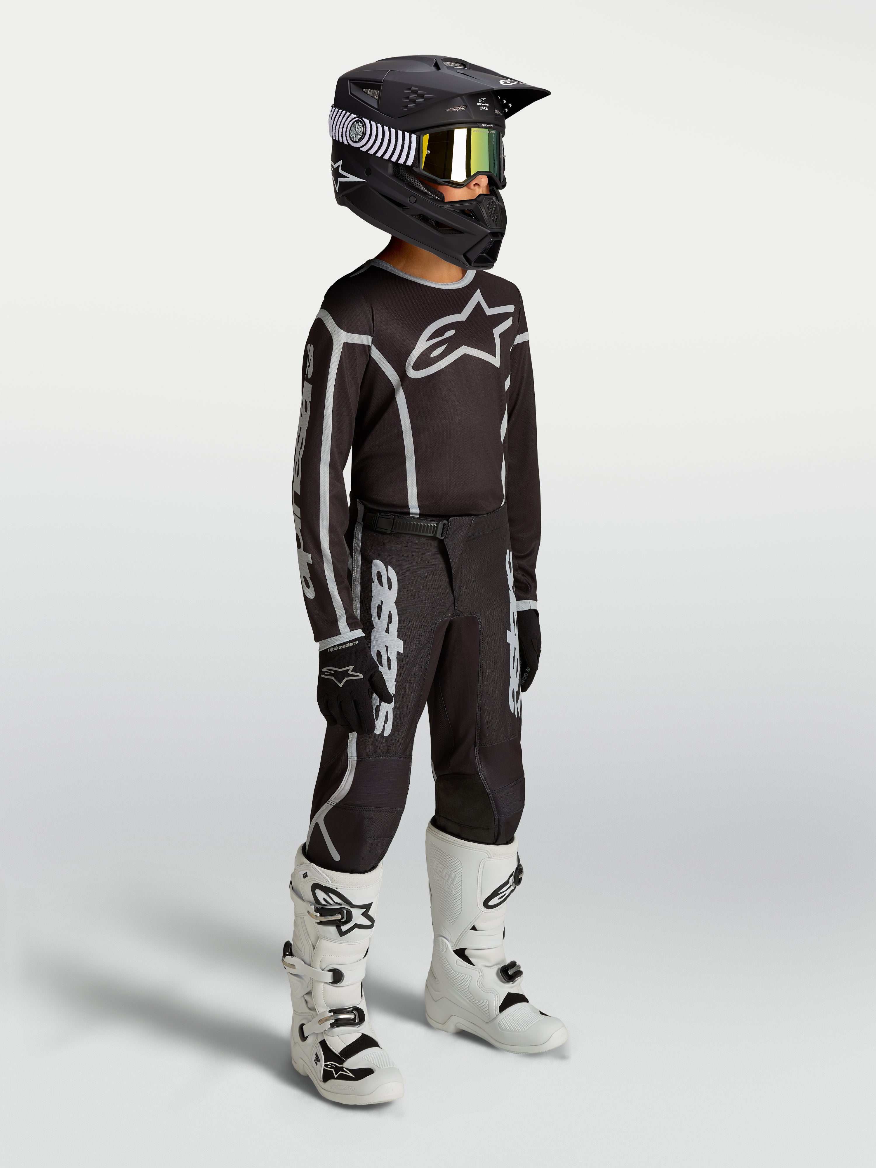 Alpinestars Jeugd Fluid Apex V2 broek, zwart en zilver motocrossuitrusting op model met jersey, helm en witte motorlaarzen.