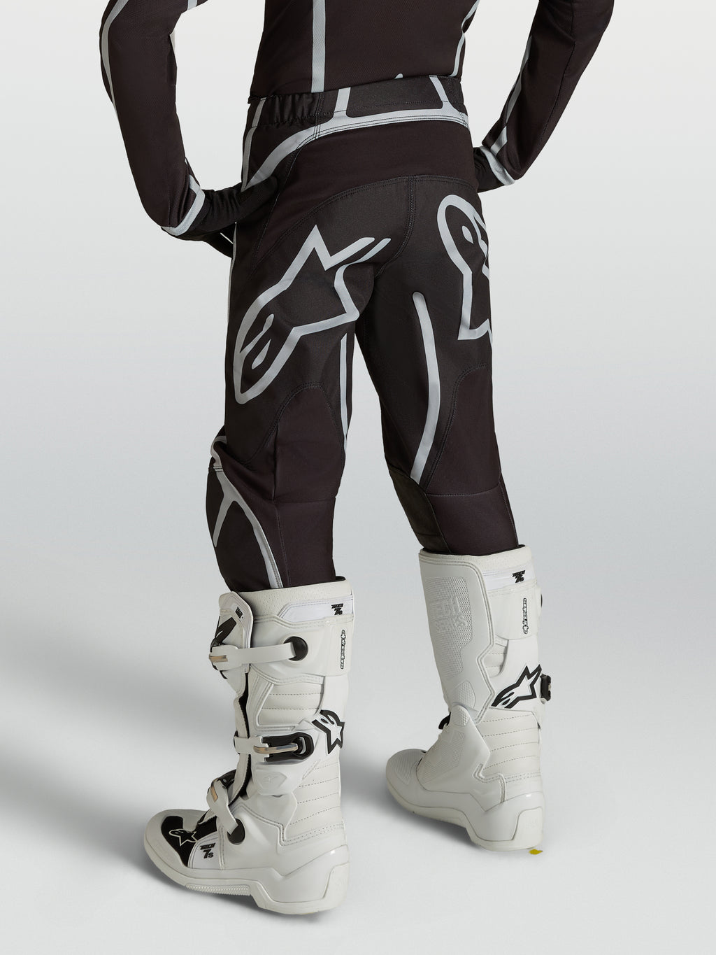 Alpinestars Jeugd Fluid Apex V2 broek, zwart-zilveren motocross-racebroek, achteraanzicht gedragen met witte motorlaarzen.