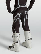 Alpinestars Jeugd Fluid Apex V2 broek, zwart-zilveren motocross-racebroek, achteraanzicht gedragen met witte motorlaarzen.