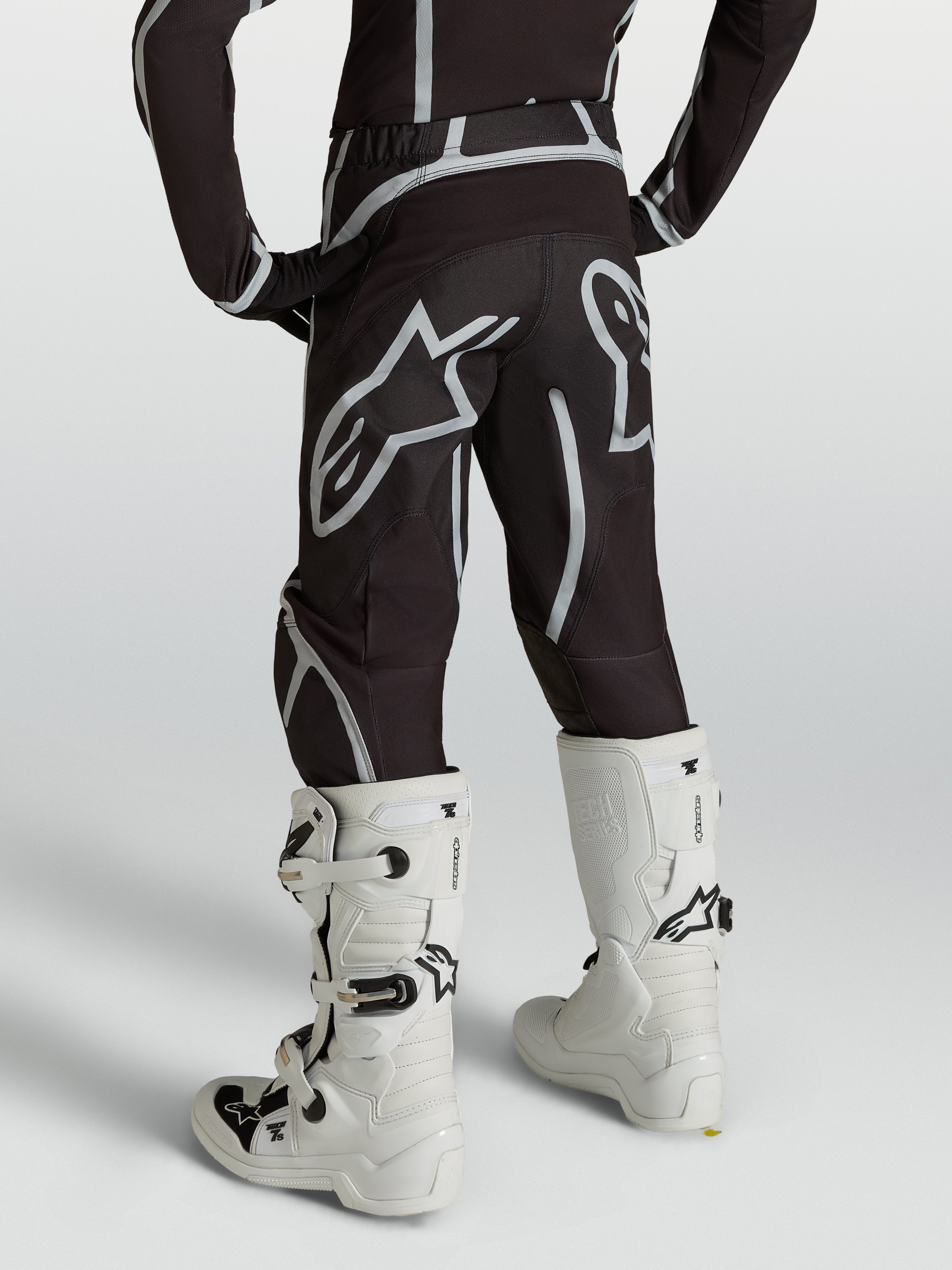 Alpinestars Jeugd Fluid Apex V2 broek, zwart-zilveren motocross-racebroek, achteraanzicht gedragen met witte motorlaarzen.