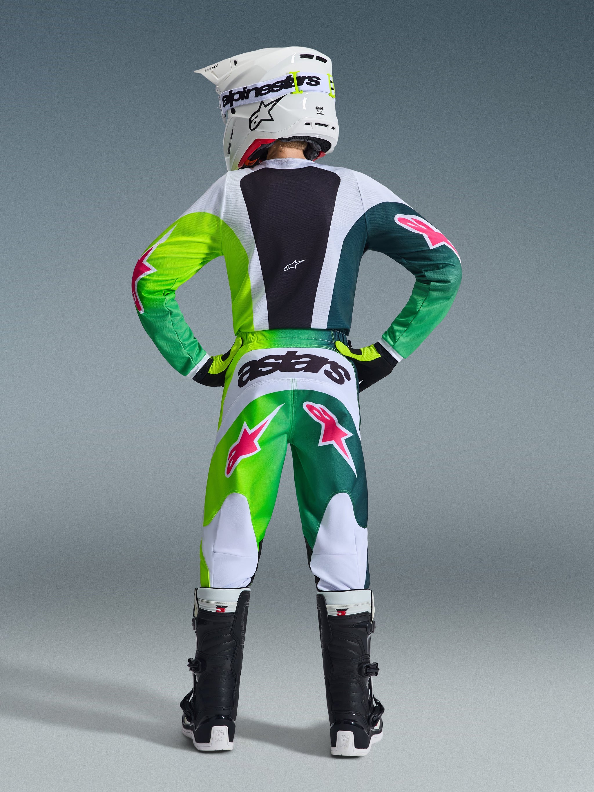 Alpinestars 2026 Jugend-Fluid Portl Hose, Motocross-Hose, Grün, Weiß und Schwarz mit pinkfarbenen Stern-Logos, Rückansicht kombiniert mit passendem Trikot und schwarzen Motocross-Stiefeln, mit markantem „astars“-Branding auf der Rückseite, technische Ausrüstung für den Jugend-Offroad-Rennsport.