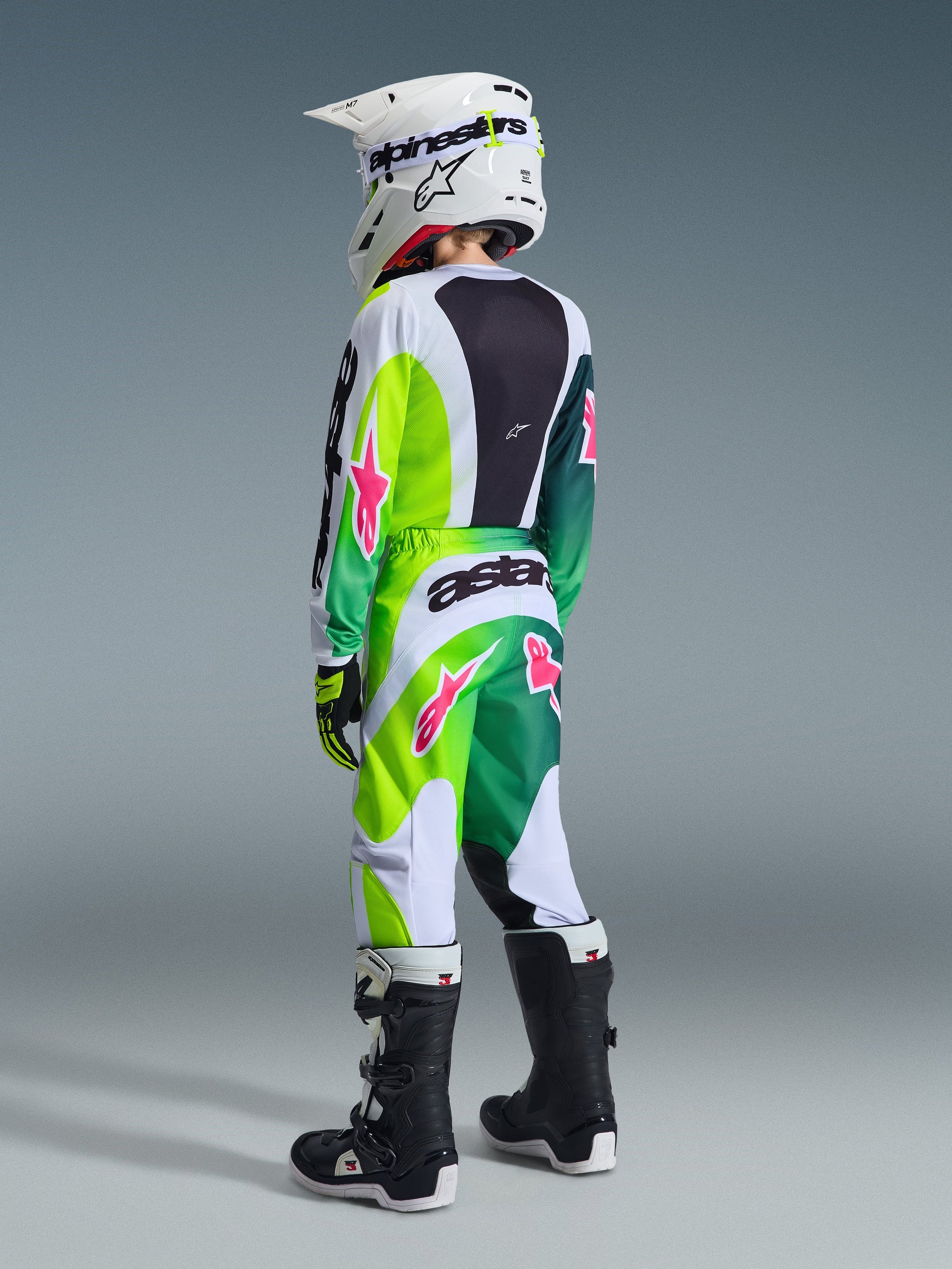 Alpinestars 2026 Youth Fluid Portl Hose, Motocross-Hose, Grün, Weiß und Schwarz mit pinkfarbenen Akzenten, Rückansicht eines jungen Fahrers in kompletter Rennausrüstung inklusive passendem Trikot, weißem Helm und schwarz-weißen Stiefeln.