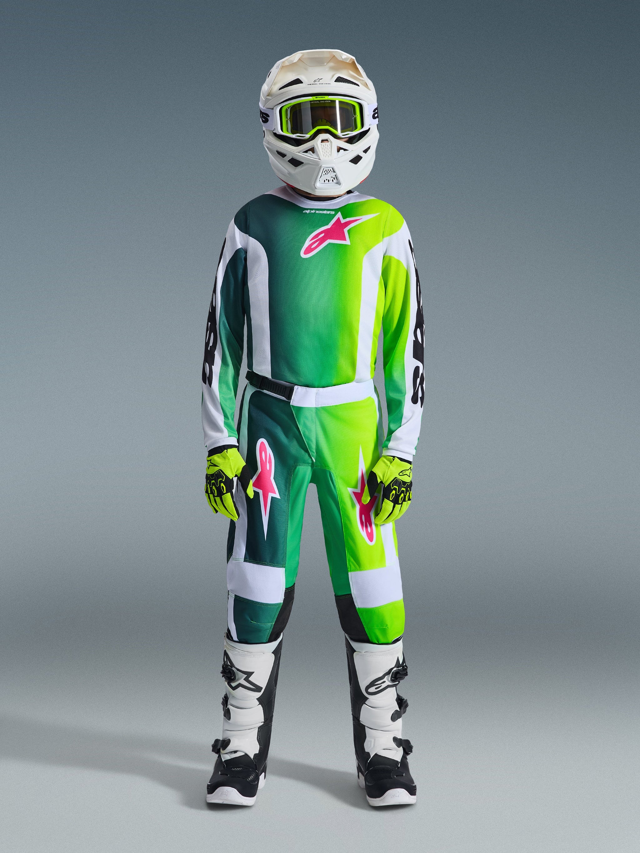 Alpinestars 2026 Youth Fluid Portl Hose, Motocross-Hose, Grün, Weiß und Schwarz, getragen von einem Jugendfahrer in kompletter Rennausrüstung inklusive passendem Trikot mit Farbverlauf, weißem Helm mit neongrüner Brille und schwarz-weißen Stiefeln, mit pinken Stern-Logos und strapazierfähigem Sport-Fit-Design.
