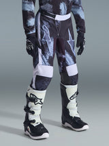 Alpinestars 2026 Youth Fluid Lahnd Pants, jeugd motorcrossbroek, Iron Camo camouflage in grijs, zwart en wit, gedragen met bijpassende jersey met lange mouwen, zwarte racehandschoenen en wit-zwarte motorcrosslaarzen, met grote sterlogo's op de dijen.