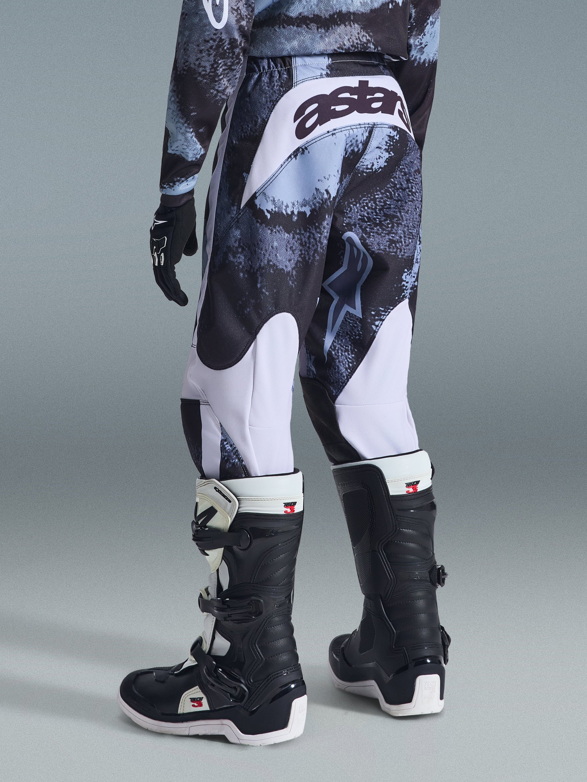 Alpinestars 2026 Youth Fluid Lahnd Pants, motorcrossbroek, Iron Camo met witte accenten, achteraanzicht met 'astars' logo op het zitvlak, gedragen met bijpassend camo shirt en zwart-witte Tech 3 motorcrosslaarzen.