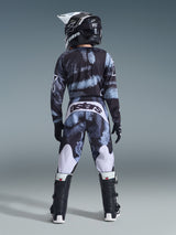 Alpinestars 2026 Youth Fluid Lahnd Pants, jeugd motorcrossbroek, Iron Camo met blauwe en grijze tinten, achteraanzicht van een rijder met een bijpassende kledingset inclusief helm en laarzen, witte accenten op de benen en 'astars' logo op de achterkant.