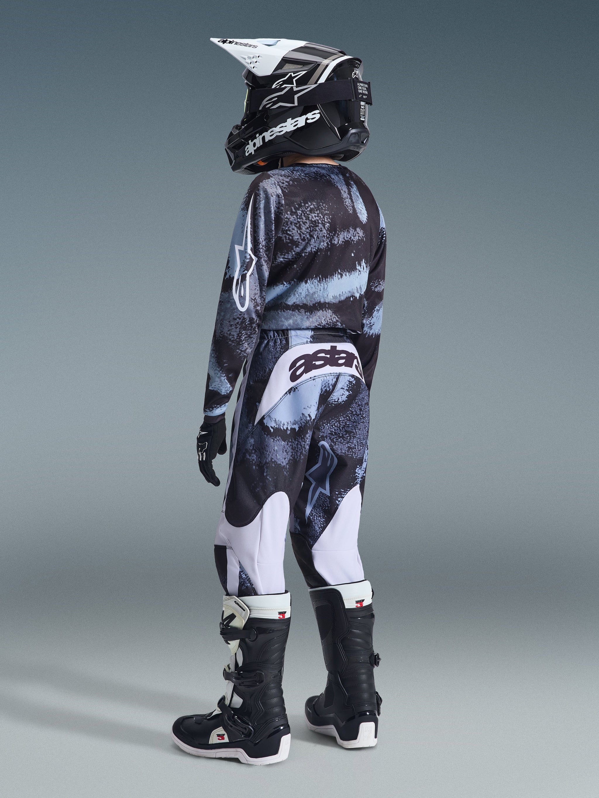 Alpinestars 2026 Youth Fluid Lahnd Pants, jeugd motorcrossbroek, Iron Camo-patroon, jonge rijder van achteren gezien in bijpassend camo-shirt, zwart-witte laarzen en zwarte helm, voorzien van witte panelen en 'astars' branding op de achterzijde, volledige uitrusting voor off-road racen