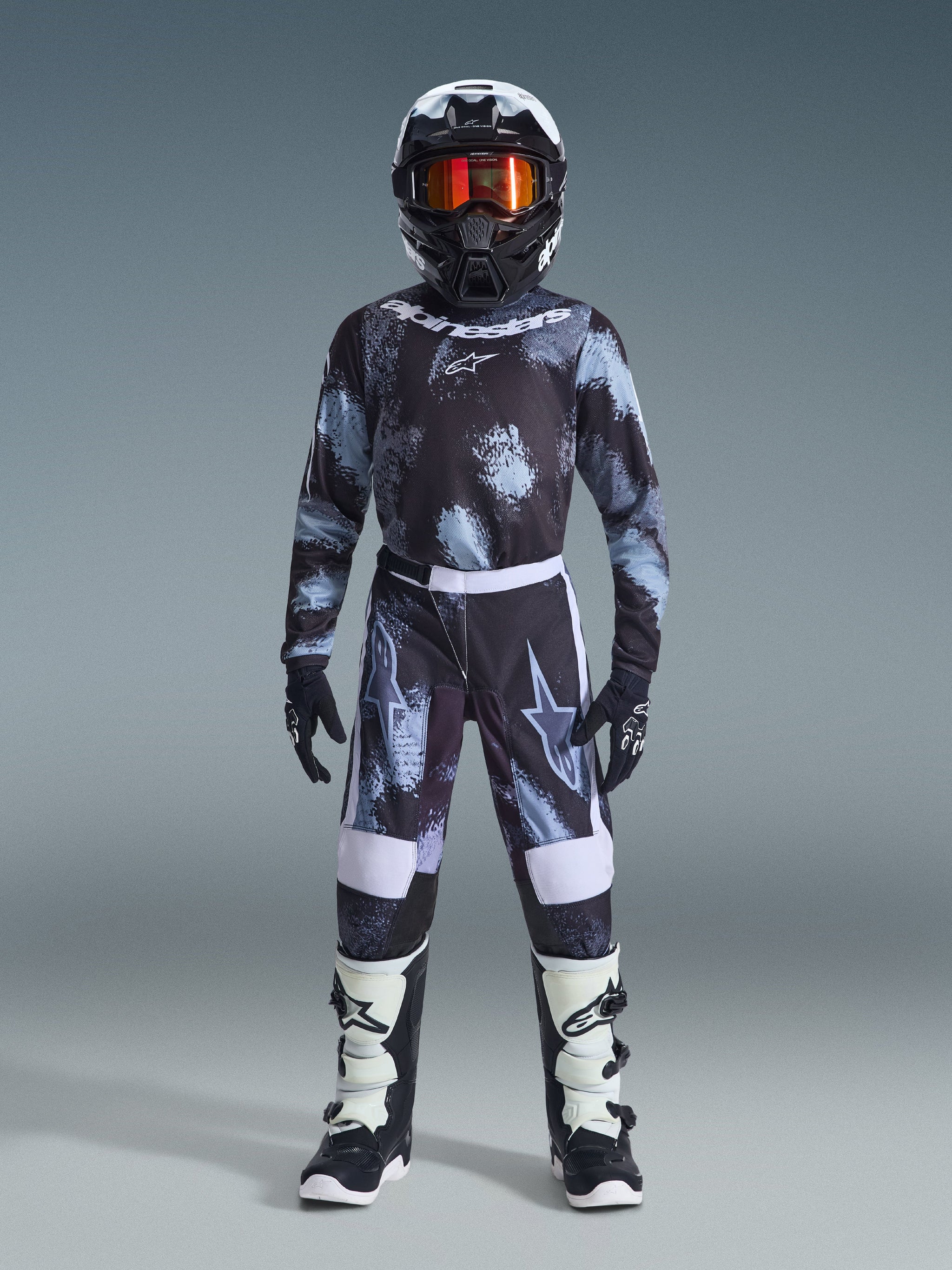 Alpinestars 2026 Youth Fluid Lahnd Pants, jeugd motorcrossbroek, Iron Camo, gedragen door een jonge rijder met bijpassend camo jersey, zwarte helm met oranje bril en wit-zwarte laarzen, voorzien van een gecoördineerd ontwerp met opvallende logo's en versterkte panelen voor off-road racing.