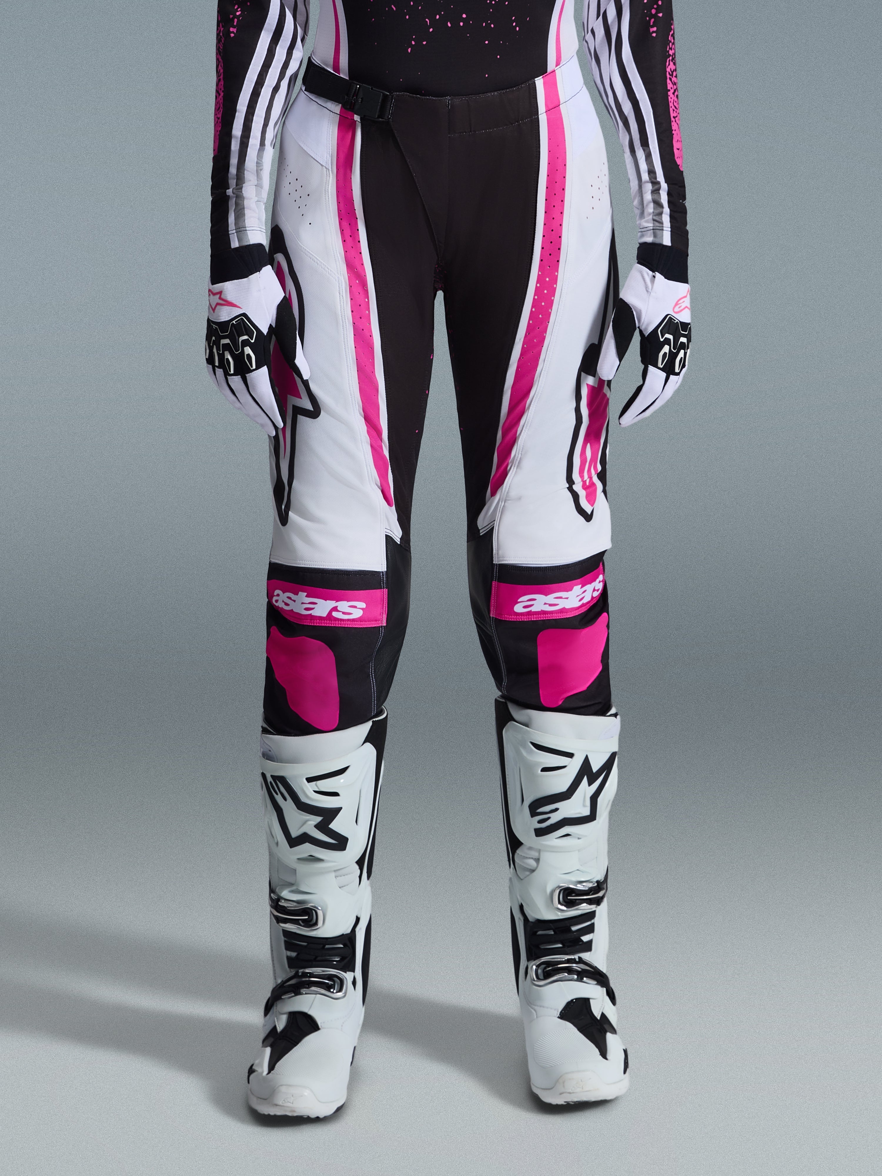 Pantalon Alpinestars Stella Techstar Nomur 2026 pour femme, pantalon de motocross, noir, blanc et fuchsia, porté avec un maillot de course, des gants assortis et des bottes de motocross blanches, doté de graphismes roses vifs et de logos astars sur les genoux, conçu pour la compétition tout-terrain de haut niveau.