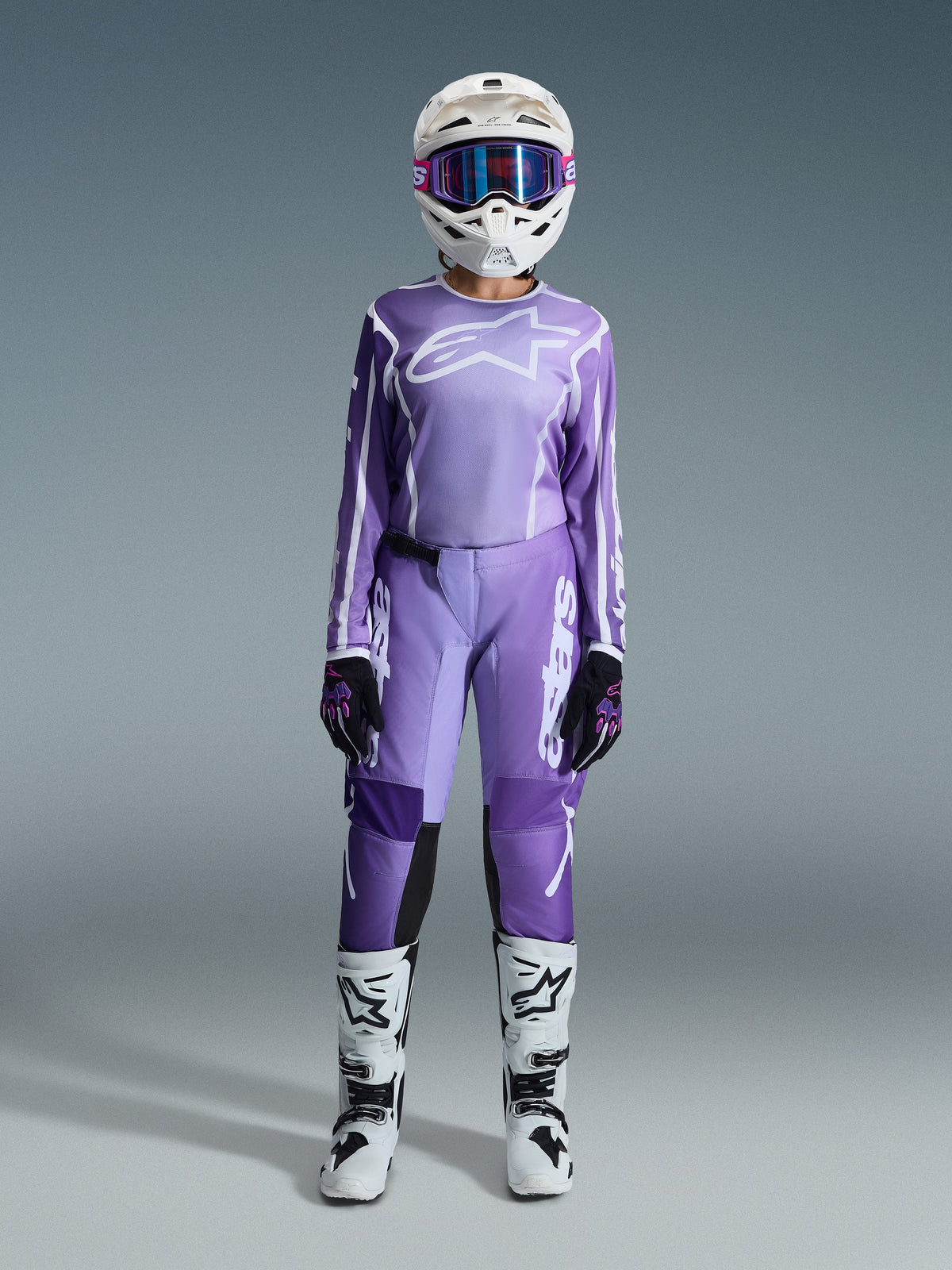 Motocrossrijder in de Alpinestars 2026 Stella Fluid Apex Gear Set voor dames, violette en witte jersey met bijpassende broek, witte helm met roze bril en witte en zwarte off-road laarzen, complete race-uitrusting voor dames motocross.