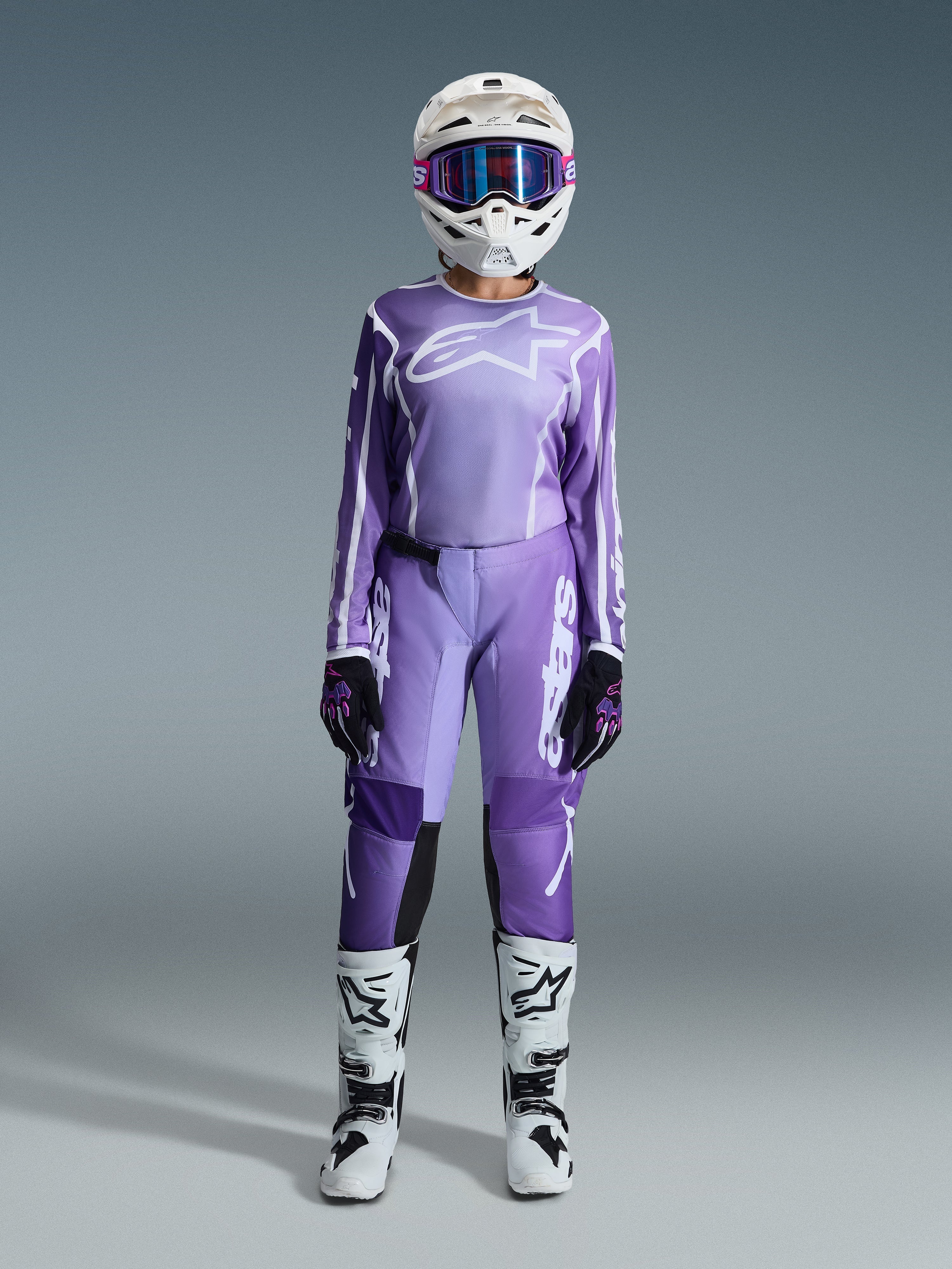 Pilote de motocross portant l'ensemble d'équipement Alpinestars 2026 Stella Fluid Apex pour femme, maillot violet et blanc avec pantalon assorti, casque blanc avec lunettes roses, et bottes tout-terrain blanches et noires, tenue de course complète pour le motocross féminin.