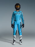 Ensemble Alpinestars 2026 Stella Fluid Apex, maillot et pantalon de motocross pour femme, bleu et noir, avec un grand logo étoile sur la poitrine, porté avec un casque noir, des lunettes teintées et des bottes blanches avec détails noirs, tenue de course tout-terrain complète