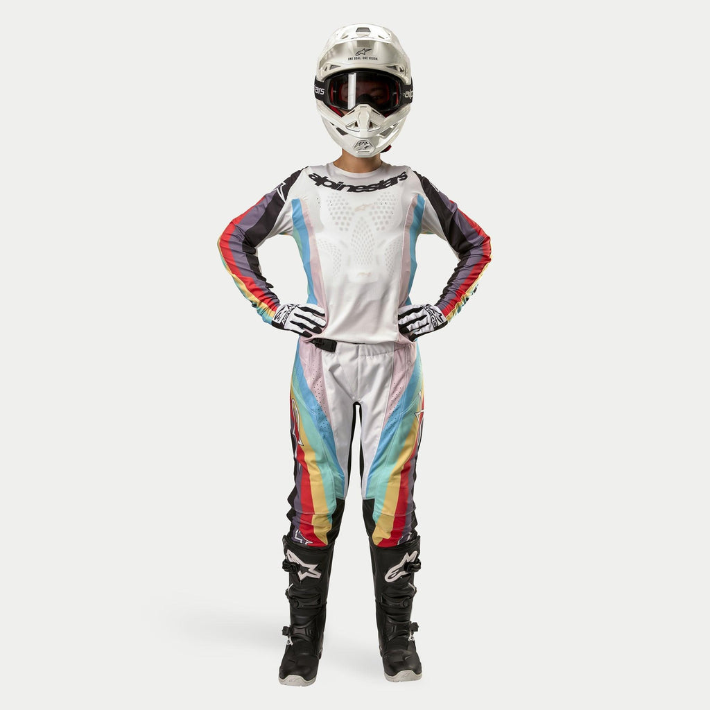 Pilote de motocross portant le pantalon Alpinestars Stella Techstar 2024 pour femme, Noir Multicolore avec base blanche et rayures rouges, jaunes et bleues, maillot à manches longues assorti, casque blanc et bottes noires, tenue de course complète pour la compétition tout-terrain
