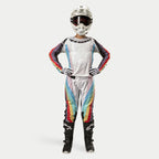Pilote de motocross portant le pantalon Alpinestars Stella Techstar 2024 pour femme, Noir Multicolore avec base blanche et rayures rouges, jaunes et bleues, maillot à manches longues assorti, casque blanc et bottes noires, tenue de course complète pour la compétition tout-terrain