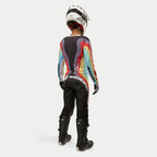 Pilote de motocross vue de dos portant le pantalon Alpinestars Stella Techstar 2024 pour femme en Noir Multicolore, avec un grand marquage blanc sur l'assise, assorti à un maillot à rayures colorées, un casque blanc et des bottes Tech 3 noires pour la course tout-terrain.