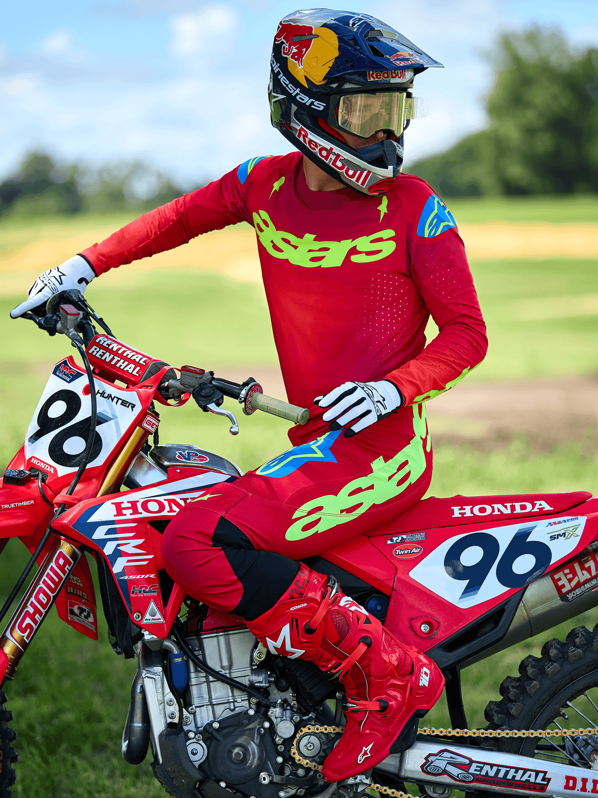 Motorcrosser draagt de Alpinestars 2025 Supertech Maker Jersey, helderrood met neongele 'astars' branding, gecombineerd met bijpassende rode broek en laarzen, witte en zwarte handschoenen, zittend op een rode Honda racemotor in een buitenomgeving