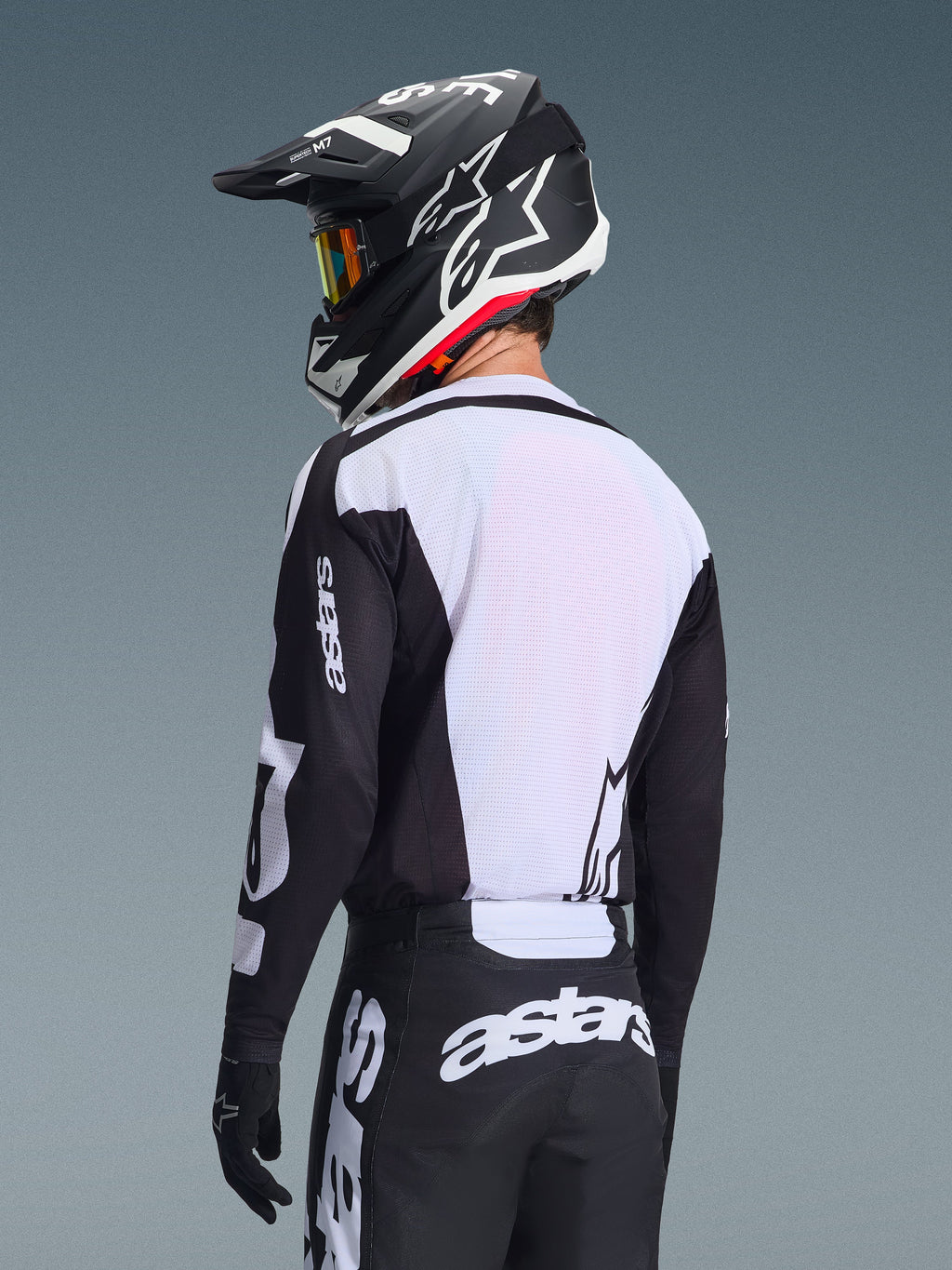 Motocross rider from behind wearing the Alpinestars 2026 Racer Air Riway Jersey, Black and White, featuring a white breathable mesh back panel and black sleeves with 'astars' branding, matching black pants with white logos, black helmet with white accents, professional off-road gear for high-temperature performance.