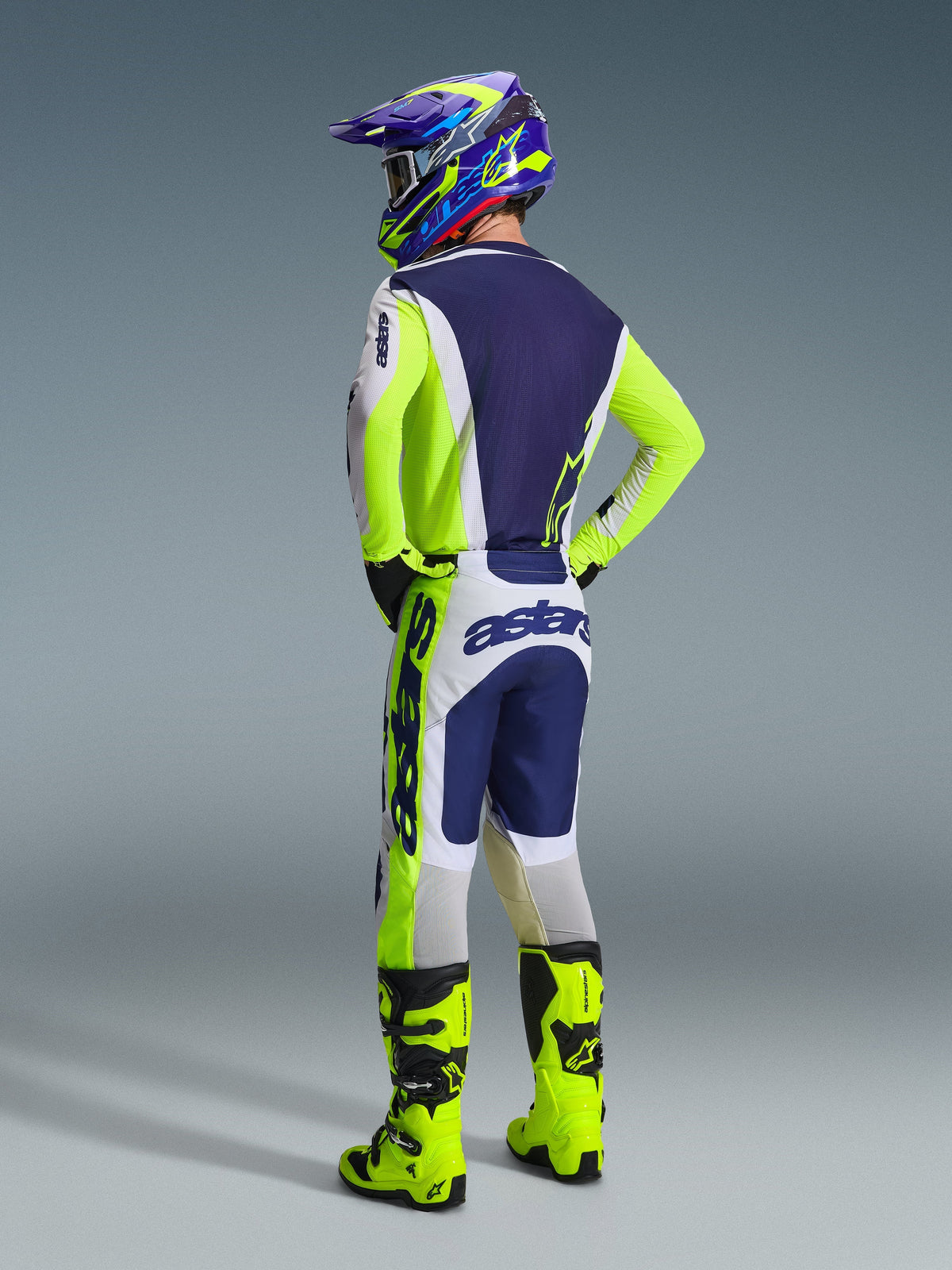 Pilote de motocross de dos portant le maillot Racer Air Riway 2026 Alpinestars en gris clair, jaune fluo et bleu, pantalon assorti avec logos imposants, bottes de motocross jaune fluo éclatant et casque bleu et jaune, ensemble complet d'équipement de course tout-terrain.