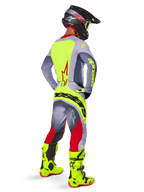Pilote de motocross vu de dos portant le maillot Alpinestars Techstar Melt 2025 en rouge et gris, avec panneau arrière jaune vif et manches grises, pantalon assorti gris et rouge avec logos jaunes, bottes de motocross jaune vif et casque noir avec lunettes jaunes, tenue de course tout-terrain coordonnée