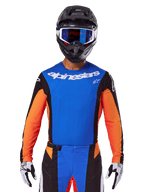 Alpinestars 2025 Techstar Melt Jersey, motocross jersey, blauw en oranje met zwarte zijpanelen, gedragen met zwarte helm en blauwe crossbril, voorzien van ademende lasergesneden perforaties op de borst, atletische racepasvorm voor off-road prestaties