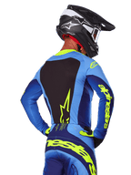 Vue arrière d'un pilote de motocross portant le maillot Alpinestars 2025 Techstar Melt bleu et jaune fluo, avec panneau dorsal noir et logo jaune vif sur les manches, associé à un pantalon de motocross assorti et un casque en carbone noir, équipement de course tout-terrain haute performance