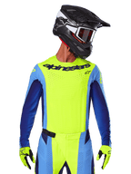 Maillot Alpinestars Techstar Melt 2025, maillot de motocross, jaune fluo et bleu, avec ventilation découpée au laser et coupe sportive de compétition, porté par un pilote avec un casque noir et des gants jaune fluo