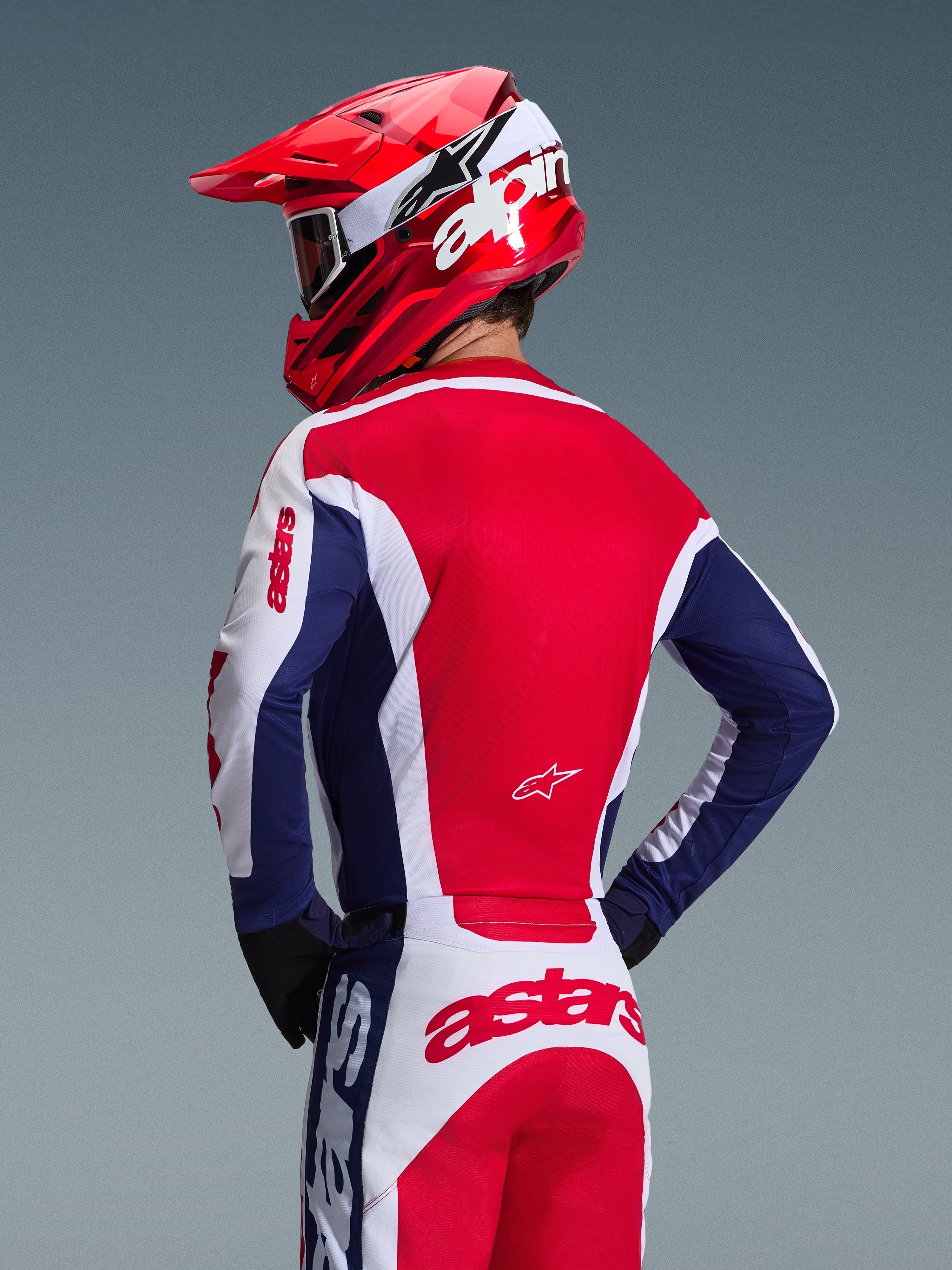 Motocross-Fahrer von hinten im Alpinestars 2026 Racer Riway Jersey, Farbkombination Weiß, Blau und Rot, passende Offroad-Hose mit auffälligem Astars-Branding, roter Helm mit weißem Alpinestars-Logo, schwarze Handschuhe, abgestimmte Motocross-Rennausrüstung