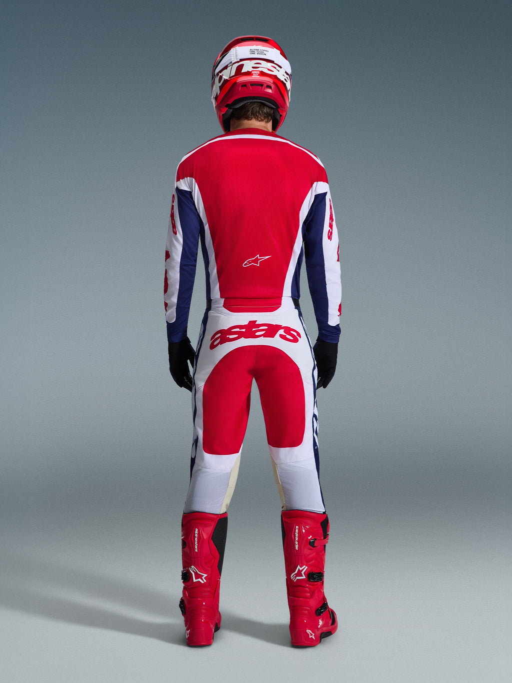 Pilote de motocross vu de dos portant l'ensemble d'équipement Racer Riway 2026, maillot rouge et bleu avec accents blancs, pantalon blanc avec empiècements rouges et logo 'astars' sur l'assise, coordonné avec un casque rouge, des bottes rouges et des gants noirs, équipement de course haute performance pour le tout-terrain
