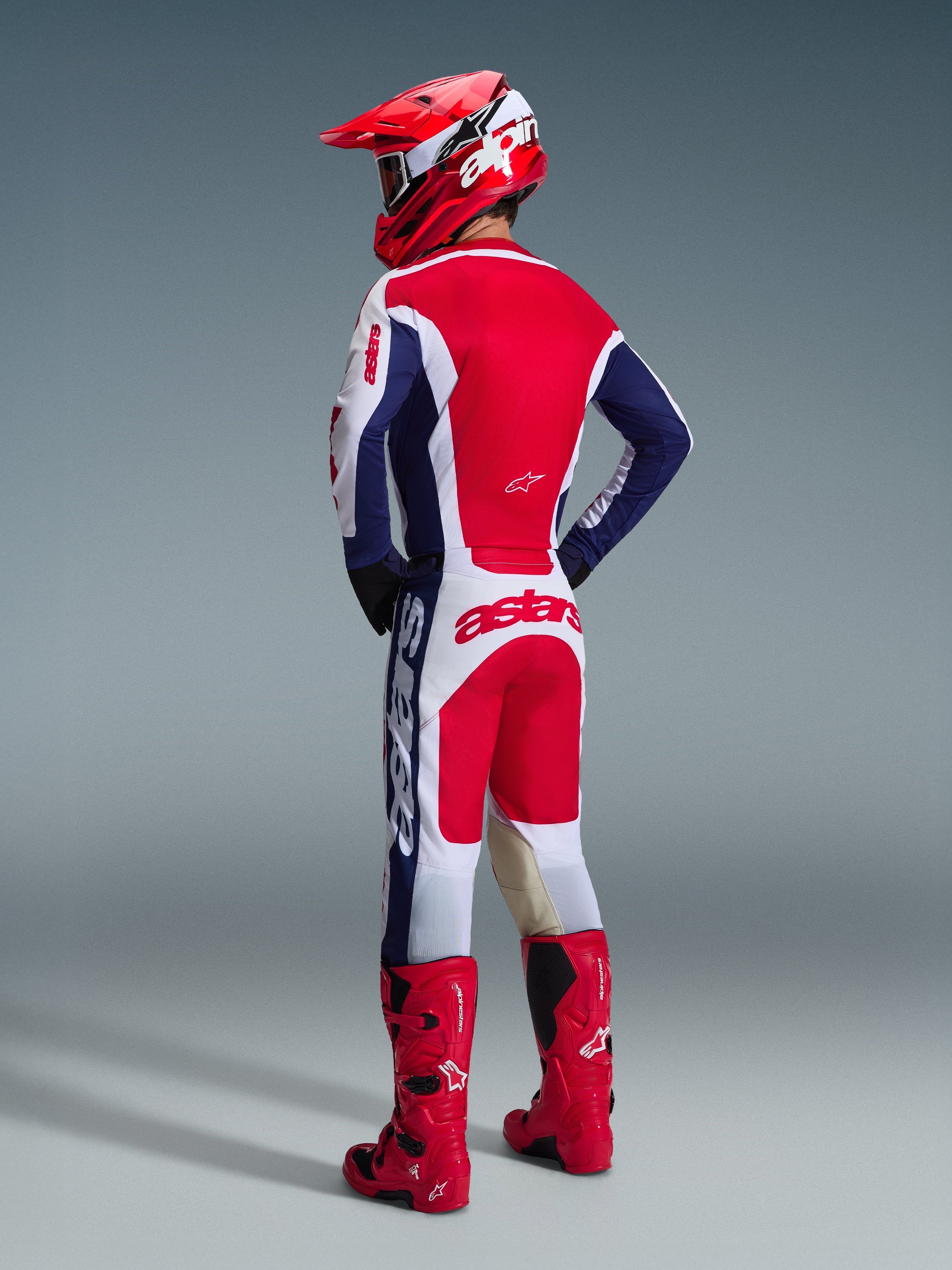 Motocross-Fahrer von hinten im Alpinestars 2026 Racer Riway Trikot in Weiß Blau Rot, passende Motocross-Hose mit „astars“-Branding auf der Sitzfläche, rot-weißer Helm und rote Motocross-Stiefel, komplette Offroad-Rennausrüstung