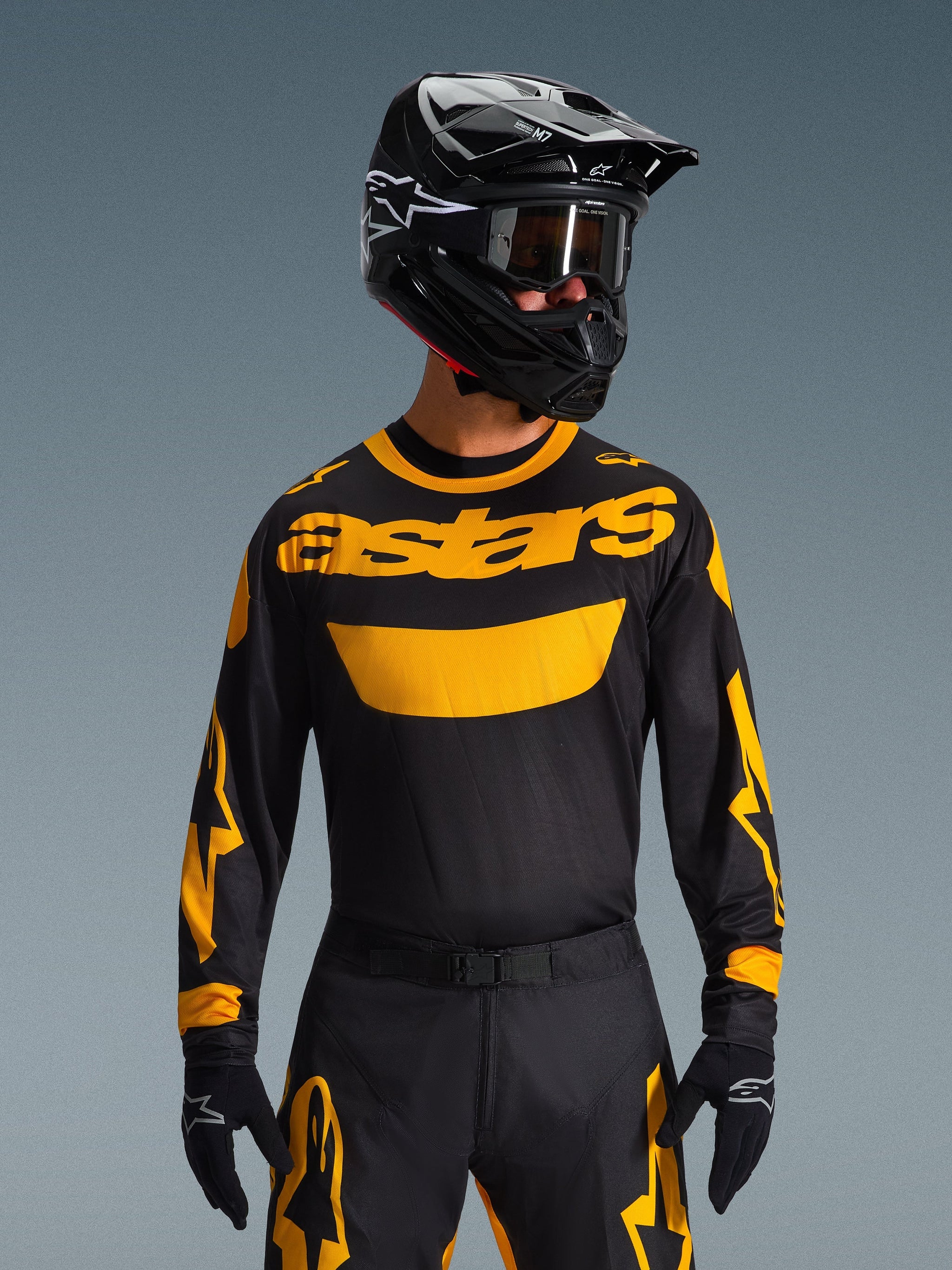 Pilote de motocross portant le maillot Alpinestars Racer Riway 2026 en jaune et noir, avec un imposant logo « astars » jaune sur la poitrine, accompagné d'un pantalon noir et jaune assorti, d'un casque noir avec lunettes et de gants noirs, équipement de course tout-terrain professionnel.