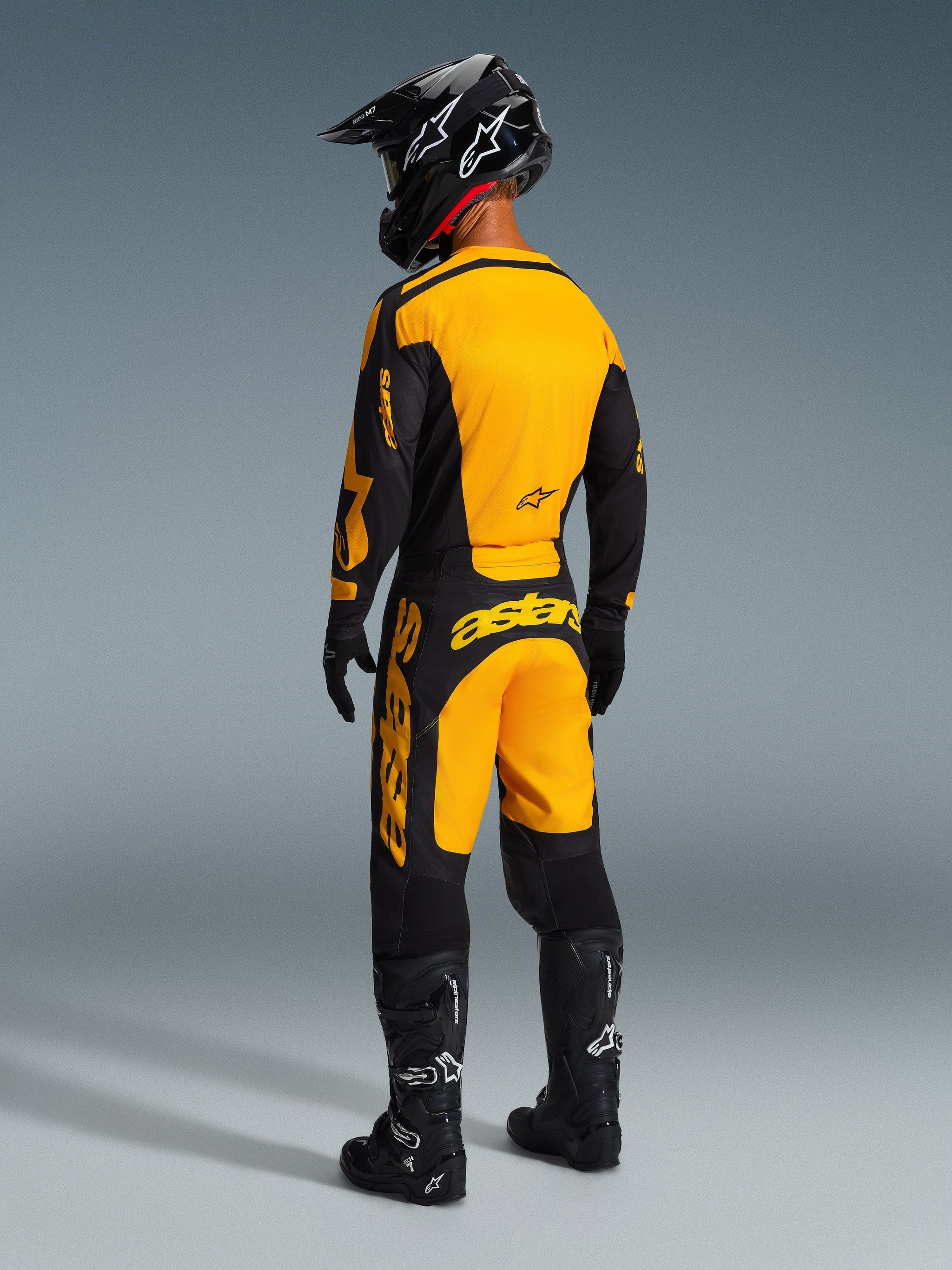 Pilote de motocross portant un équipement de course complet, maillot Alpinestars Racer Riway 2026 jaune et noir, pantalon jaune et noir assorti, casque noir, gants noirs et bottes noires, vue arrière d'une tenue tout-terrain coordonnée