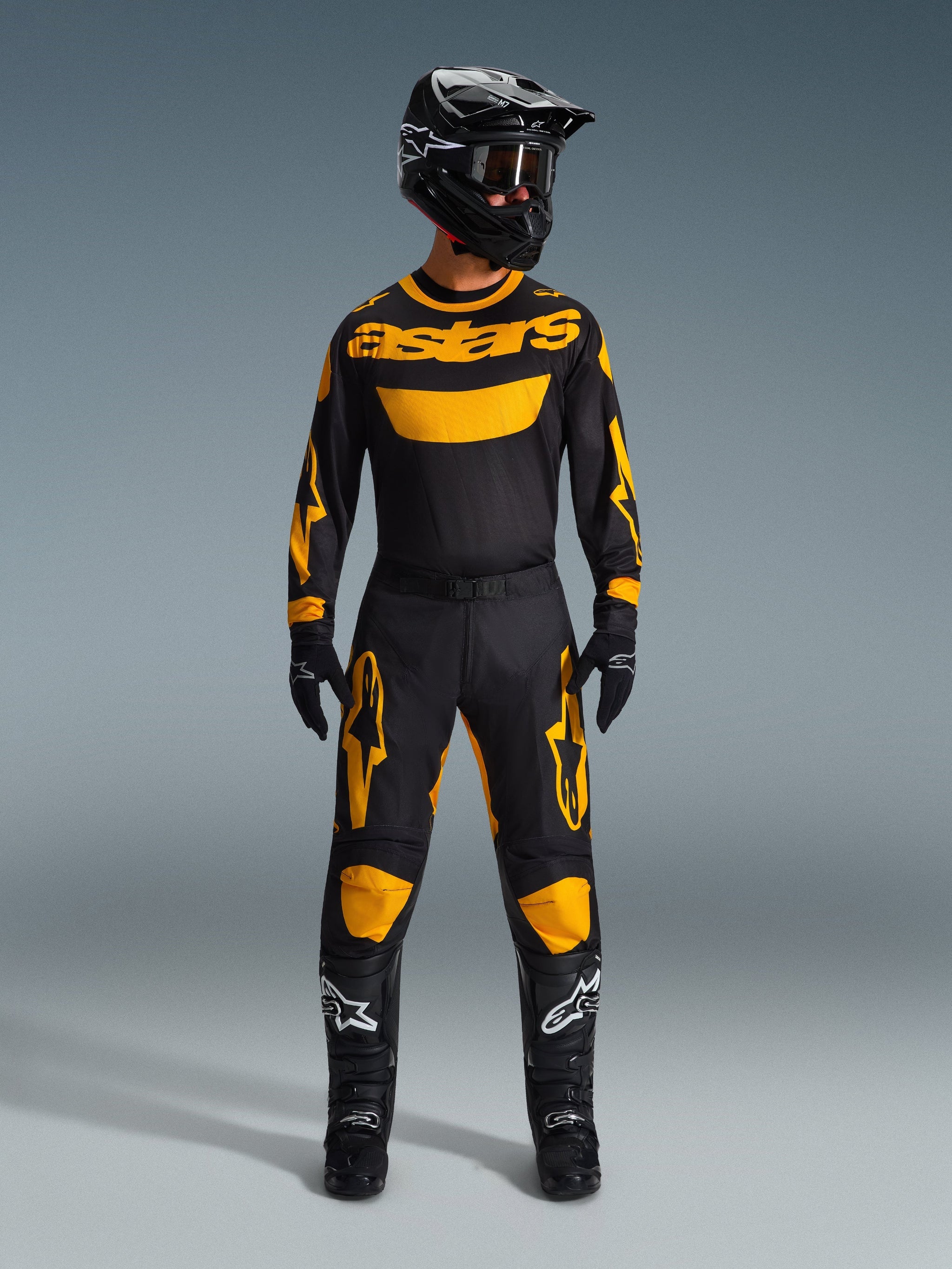 Pilote de motocross portant le maillot Racer Riway 2026 jaune et noir, coordonné avec un pantalon assorti, un casque noir et des bottes noires avec des logos Alpinestars blancs, équipement complet pour la course tout-terrain.