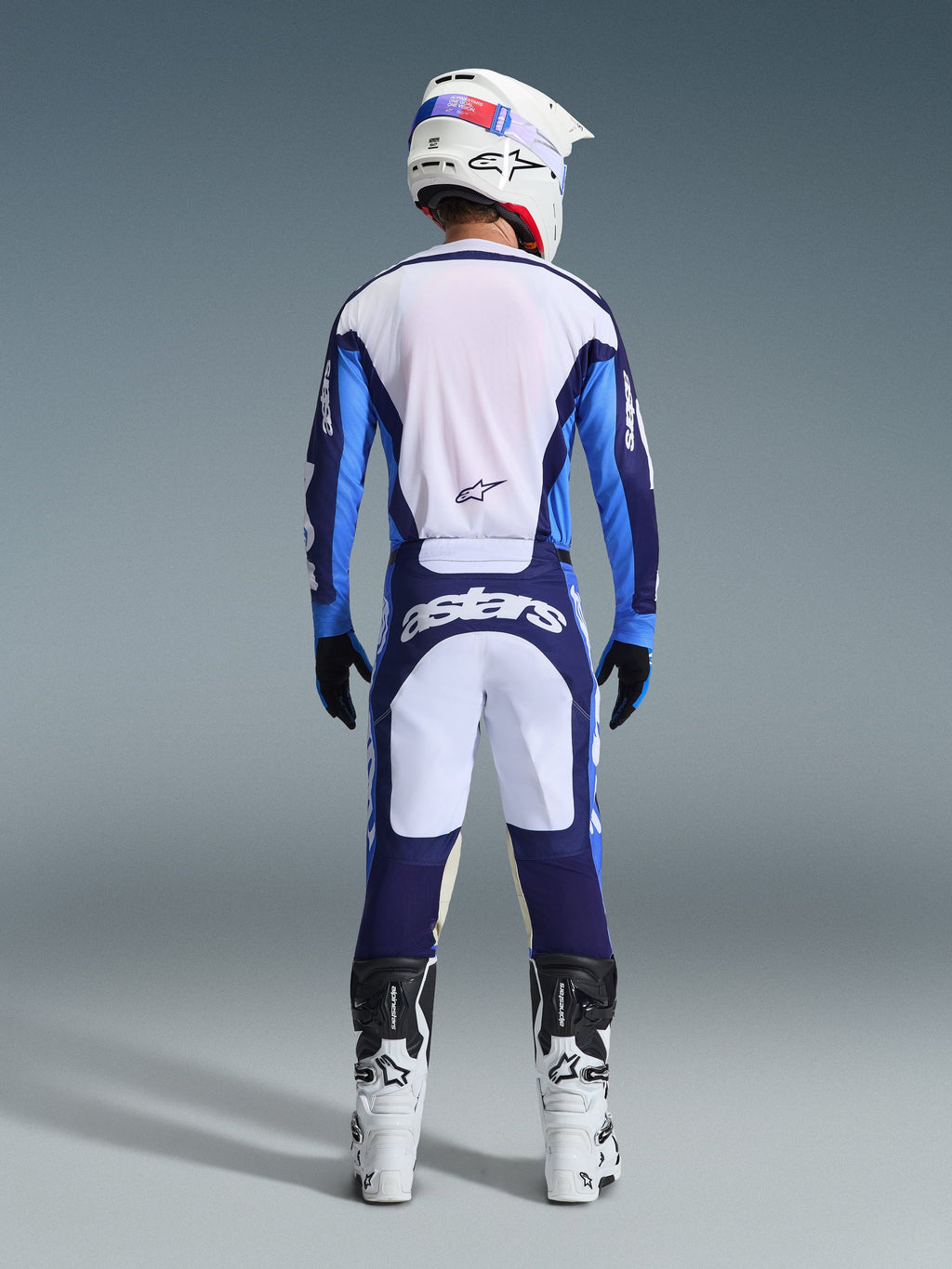 Rückansicht eines Motocross-Fahrers mit dem Alpinestars 2026 Racer Riway Trikot und der Hose in Ucla Blau und Weiß, ergänzt durch einen weißen Helm, schwarze Handschuhe und weiße Motocross-Stiefel, Hochleistungsausrüstung für Offroad-Rennen