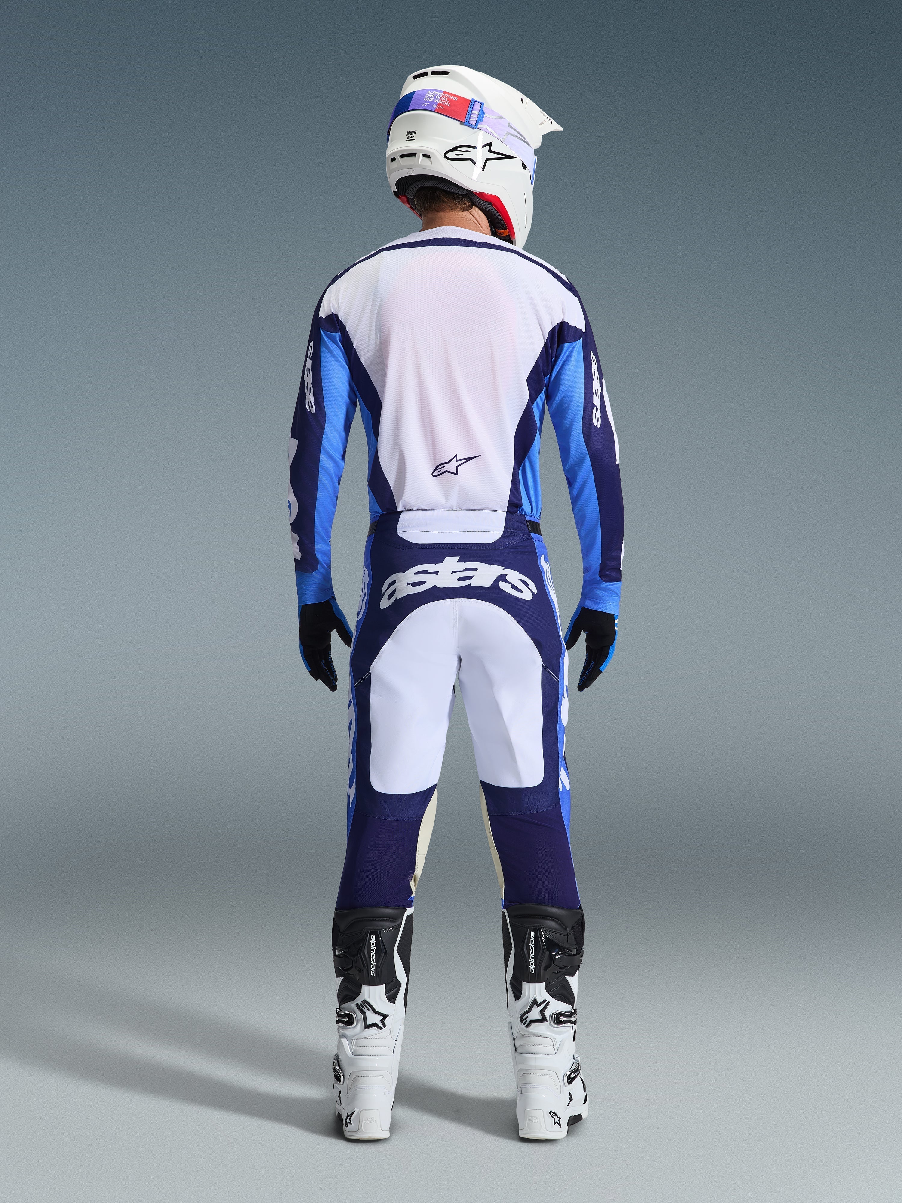 Motocross-Fahrer von hinten im Alpinestars 2026 Racer Riway Trikot in Ucla-Blau und Weiß, passende Motocross-Hose mit weißen und dunkelblauen Einsätzen und Astars-Branding, weiße und schwarze Stiefel, weißer Helm, komplettes abgestimmtes Rennoutfit für Offroad-Performance.