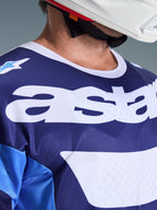 Alpinestars 2026 Racer Riway Jersey, Motocross-Trikot, Ucla-Blau und Weiß, Nahaufnahme des Brustbereichs mit großem weißem „astars“-Branding, weißem Kragen und feuchtigkeitsableitender Mesh-Struktur, getragen mit einem weißen Helm