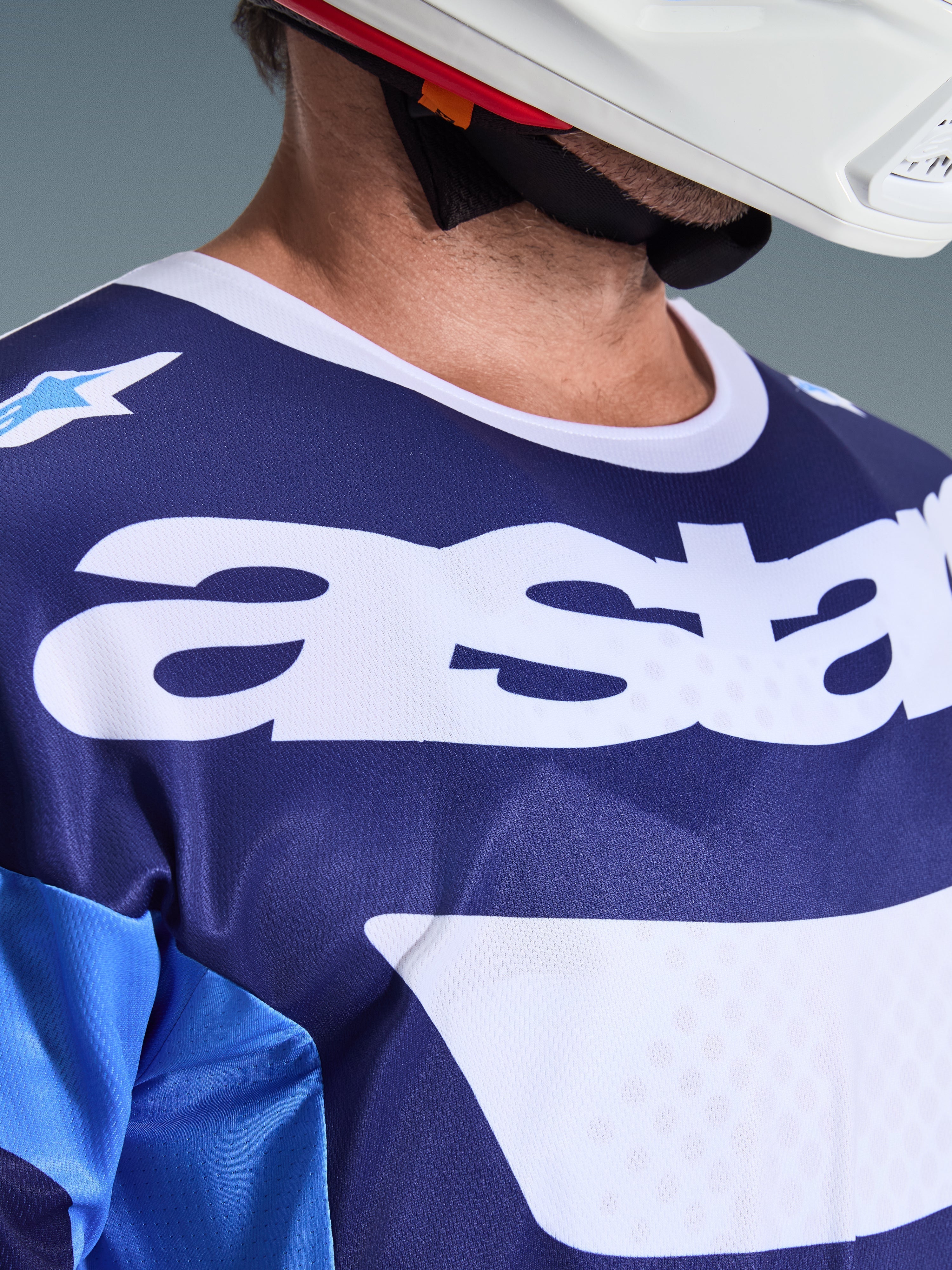 Nahaufnahme des Alpinestars 2026 Racer Riway Trikots in Ucla Blue und Weiß, mit großem weißem „astars“-Logo auf der Brust, weißem Kragen und atmungsaktiver Mesh-Gewebe-Struktur, getragen mit einem weißen Motocross-Helm
