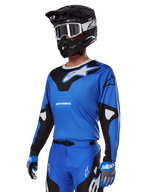 Alpinestars 2025 Racer Veil Gear Set, blauwe motorcross jersey met zwart-witte graphics, gedragen met bijpassende broek, zwarte helm, witte crossbril en blauwe handschoenen, technische kleding voor off-road racen