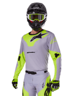 Pilote de motocross portant l'ensemble d'équipement Alpinestars Racer Veil 2025 en gris et vert fluo, maillot et pantalon gris clair avec des empiècements jaune-vert fluo vifs et des logos Alpinestars noirs, coordonné avec des gants assortis et un casque noir avec des lunettes vert fluo, tenue de course tout-terrain professionnelle.