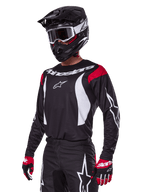 Motocross-Fahrer im Alpinestars 2025 Fluid Haul Ausrüstungsset, schwarzes Trikot mit roten Schultereinsätzen und weißen Grafiken, passende schwarze Hose mit weißen Stern-Logos, rote und schwarze Handschuhe, schwarzer Helm mit weißen Akzenten und Schutzbrille, abgestimmte Motocross-Bekleidung.