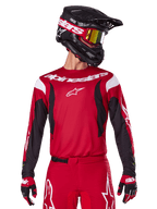 Alpinestars 2025 Fluid Haul Trikot, Motocross-Trikot, Hellrot und Schwarz, getragen als Teil eines kompletten Ausrüstungssets mit passender Hose und Handschuhen, schwarzer Helm mit gelb getönter Brille, weißes Branding und Logos, Hochleistungs-Offroad-Rennbekleidung