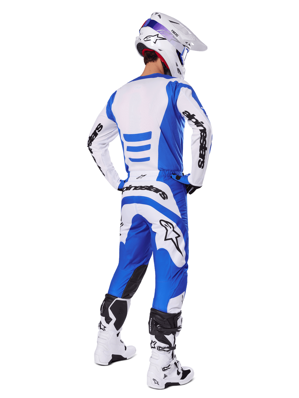 Pilote de motocross de dos portant l'ensemble d'équipement Alpinestars Fluid Haul 2025, maillot bleu et blanc avec pantalon assorti, casque blanc avec accents bleus et bottes de course blanches et noires, tenue de course tout-terrain coordonnée